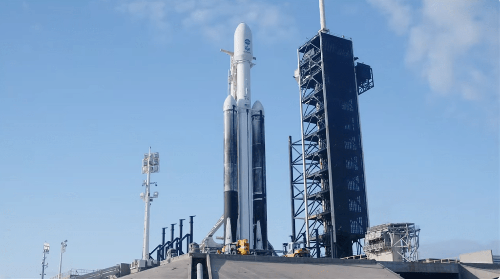 Europa Clipper sits atop a rocket at Launch Pad 39A at Kenned Space Center. The spacecraft is enclosed in a white fairing with the blue NASA logo, and the Europa Clipper mission identifier visible. The main rocket is white. The two booster rockets have white nose cones, but the bodies are charred. The SpaceX Fixed Service Structure sits to the right side of the rocket. It's a tall, gray metal structure with a white stick on top. At least two vehicles are on the ground near the rocket. Light poles are to the left of the rocket.